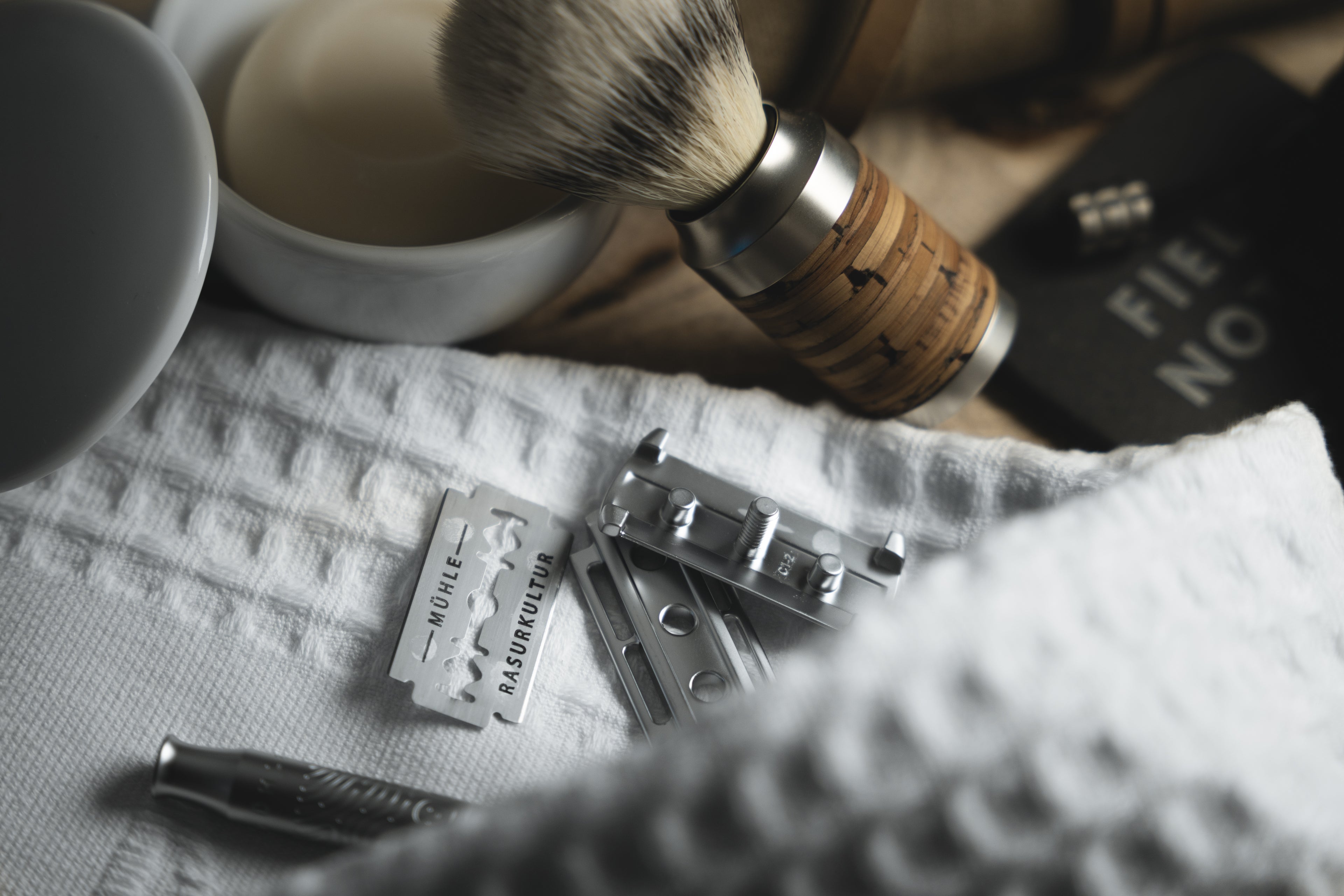 Shaving set with a shaving brush with a wooden handle, a deconstructed safety razor, and a shaving bowl place on a pique shaving towel.
