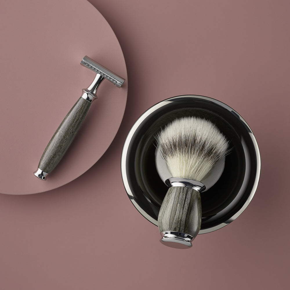 Shaving set with safety razor and a shaving brush both with a grey wooden-handled placed inside a black shaving bowl, on a pink background.