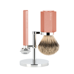 A three-piece shaving set consisting of a silvertip badger brush and a safety razor with a pink handle on a metal stand, all displayed against a white background.