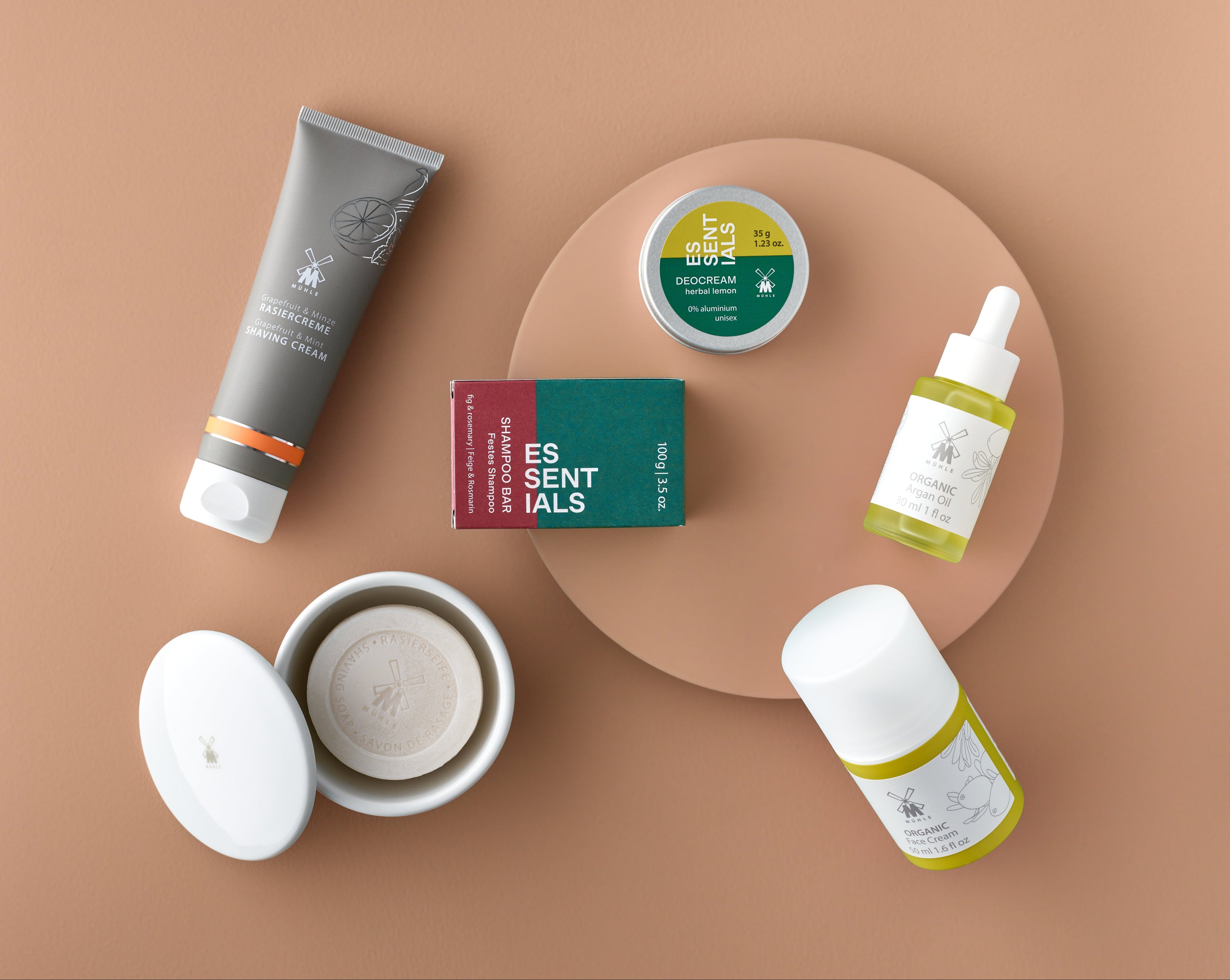 Muhle skincare and grooming products, including a tube shaving cream, soap bar, deodorant, and bottle argan oil arranged on a beige background.