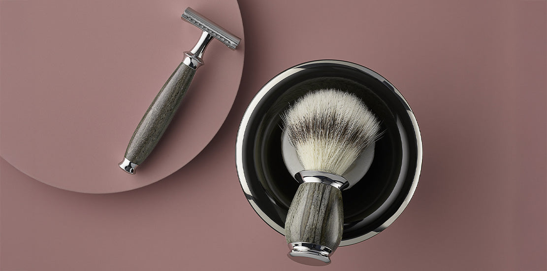 safety razor and a shaving brush with wooden handles, placed beside a metallic shaving bowl on a muted pink background.